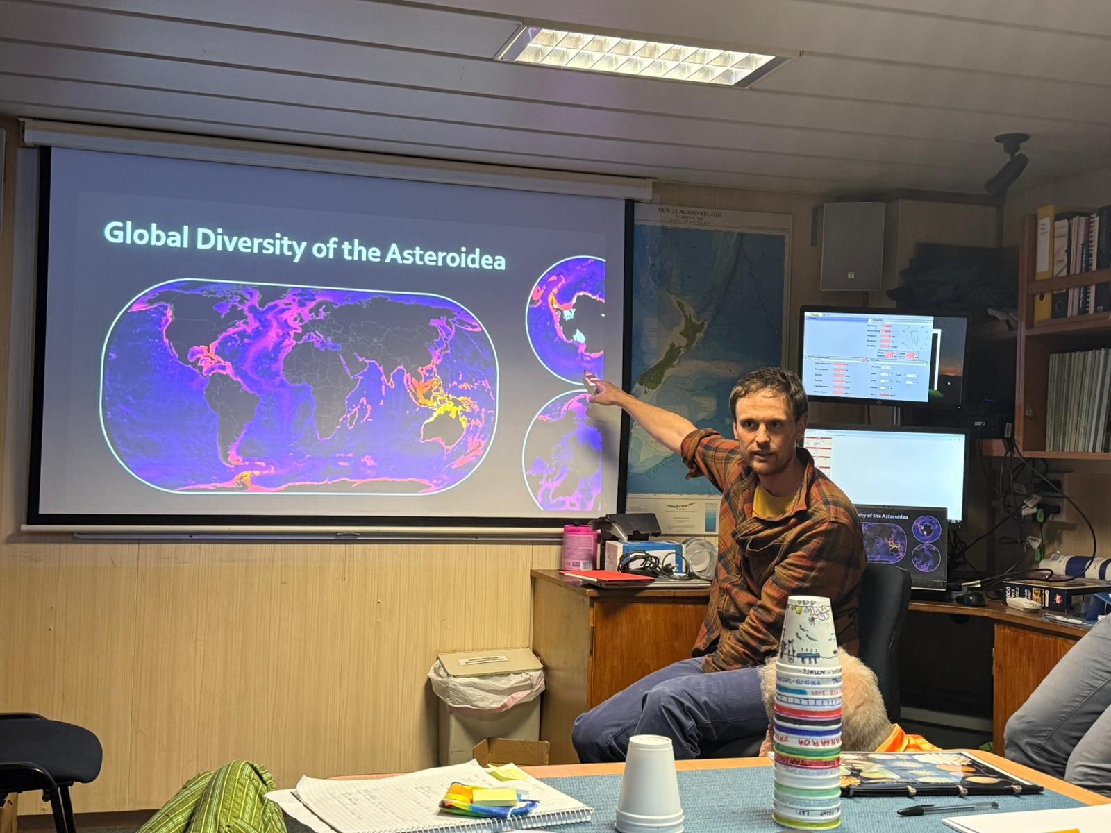 someone giving a talk in a low-ceilinged room - they are pointing at the screen which has a world map on it.   What you can't see because its a photo is the room is slowly rocking back n forth in the swell as the ship heads south. IN the foreground there are a pile of coloured cups.