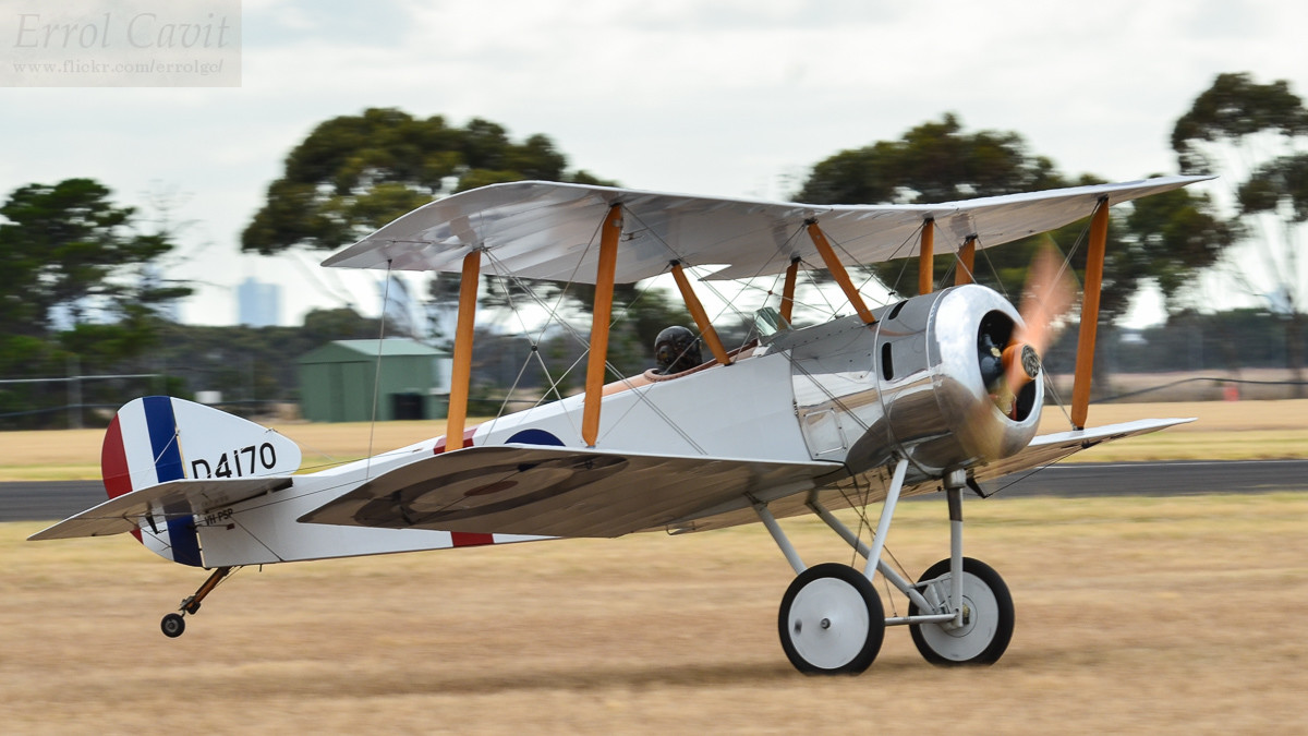 White biplane fighter just about to lift off from a grass field. The grass is brown, with sparse trees beyond and harsh sky above.