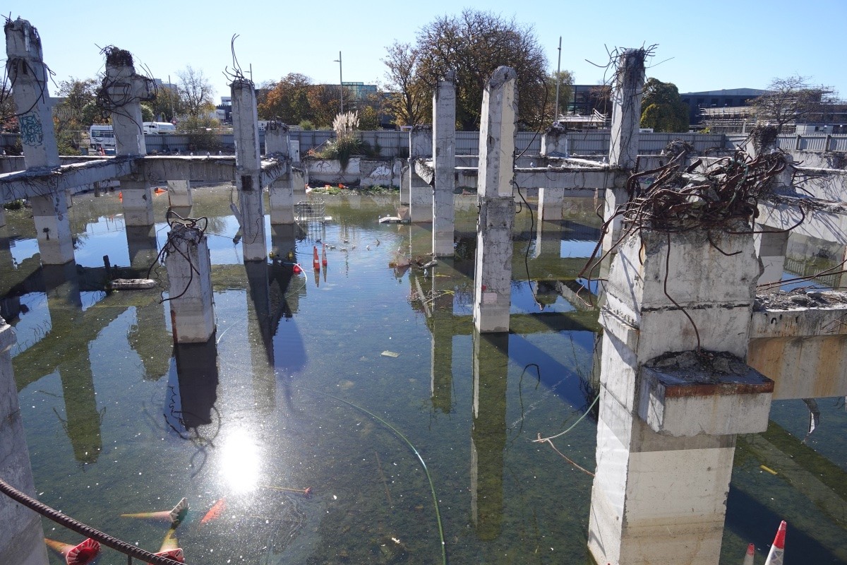 A photo from 2021 of the flooded foundations of demolished large city building. Some of the supporting concrete pillars still stand, and the basement has filled with rainwater, creating a series of small islands for seagulls to roost and nest on.
