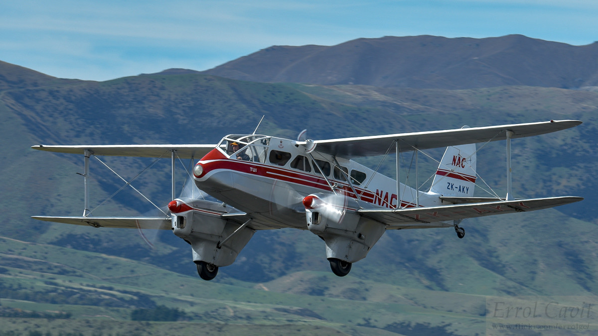 Small biplane airliner in front of steep hills, taken from front left quarter. It is silver with white upper fuselage and tail, red cheat lines between.