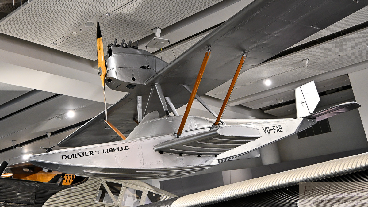 Small silver metal flying boat hung in a museum. Its engine is fitting on the parasol wing, above the cockpit. It also has significant sponsons below the wooden wing struts attached to the fuselage.