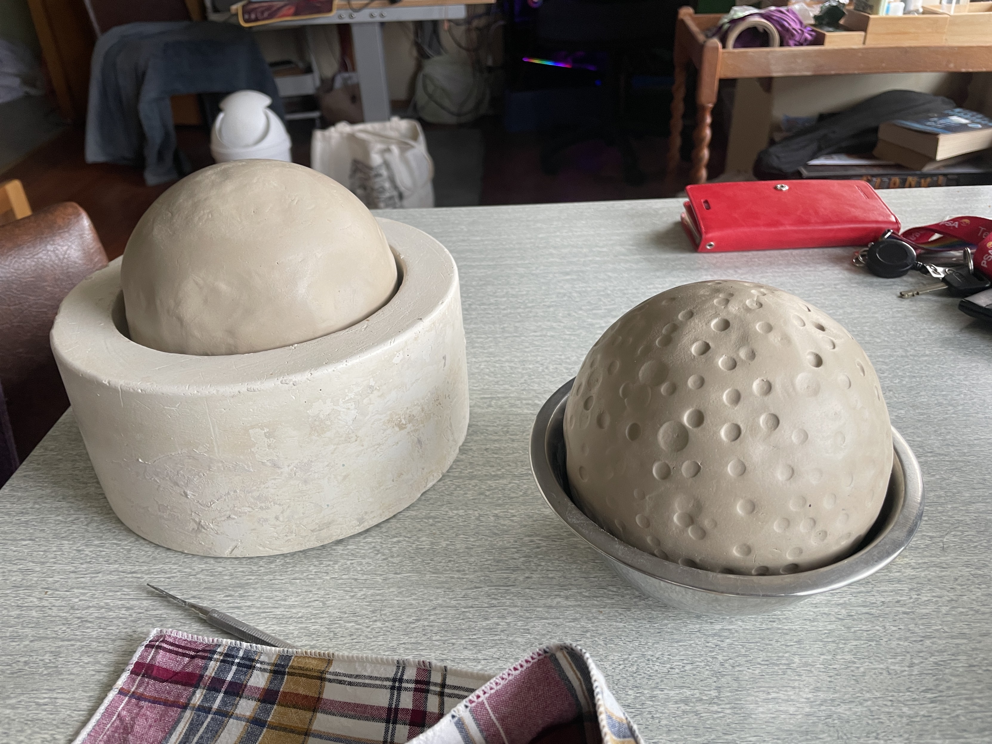 two clay orbs sitting in vessels that they have been shaped in. one is smooth beige clay in a white ceramic bowl, the other is pockmarked suggesting a lunar surface and is resting in a round steel bowl