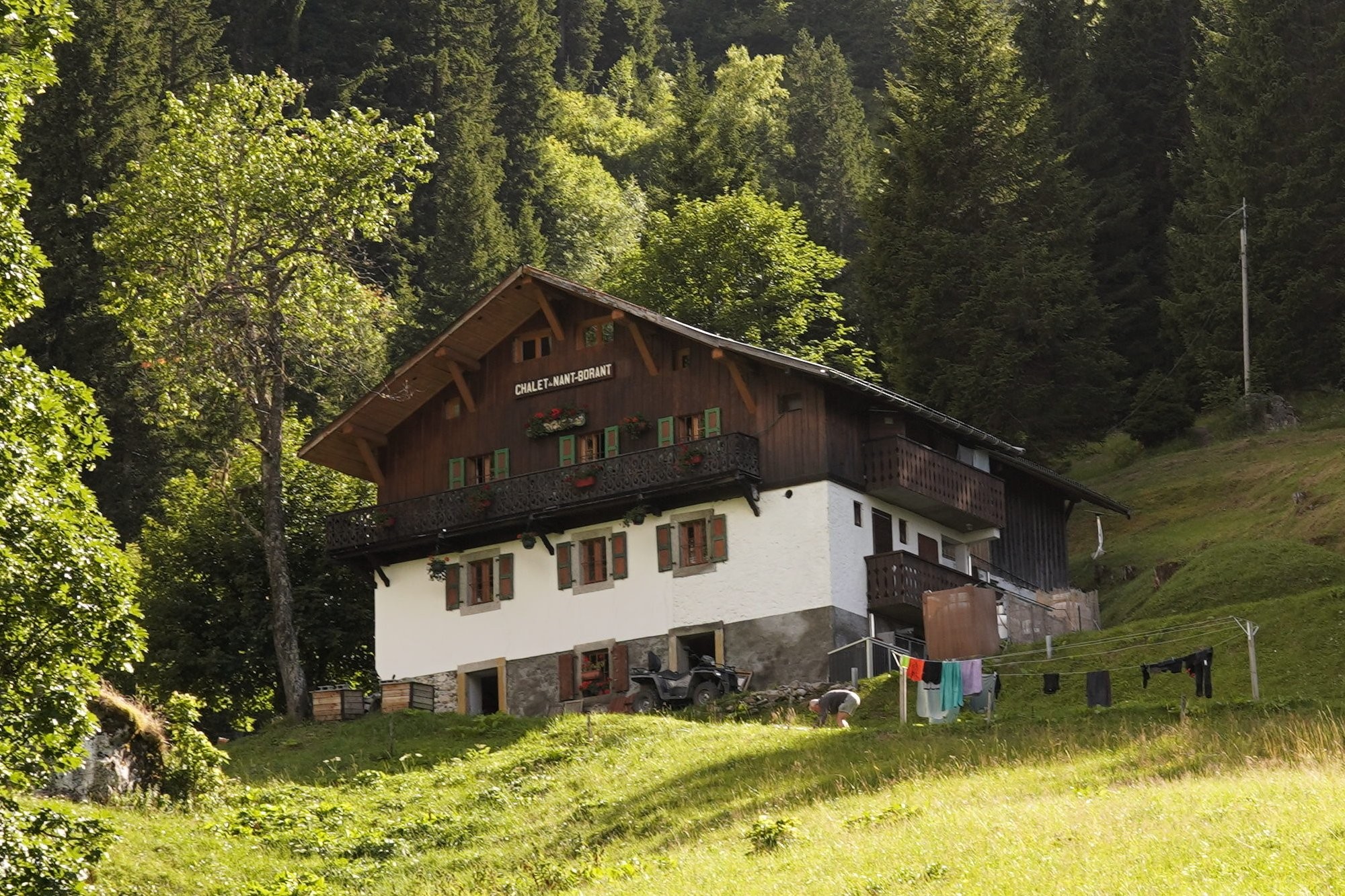 Another Alpine looking building, the Chalet Nant Borrant has wood panelling on top of stone cladding. There is a 4x4 outside the door and people are hanging various clothes out to dry. It's on some grassy slopes next to a European forest.