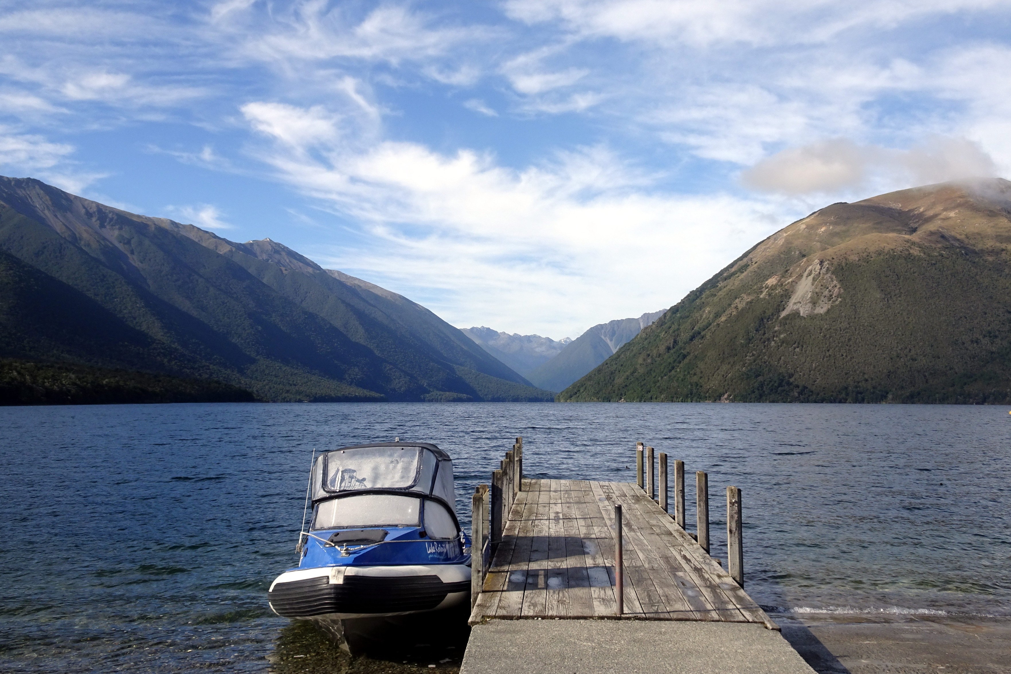 A boat is tied up at the pier on Lake Rotoiti. The Travers Valley is visible in the distance, as a V shape from here, between two ridges, with more mountains visible in the distance. The sky is partly cloudy.