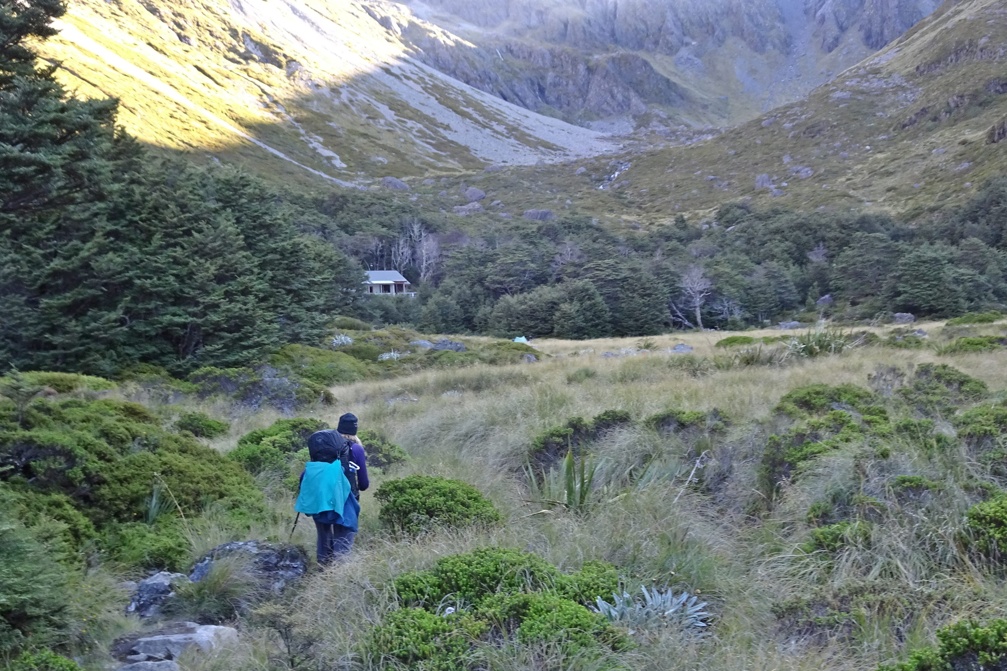 A person walks across a meadow towards a hut in the distance. Scree fields further off.