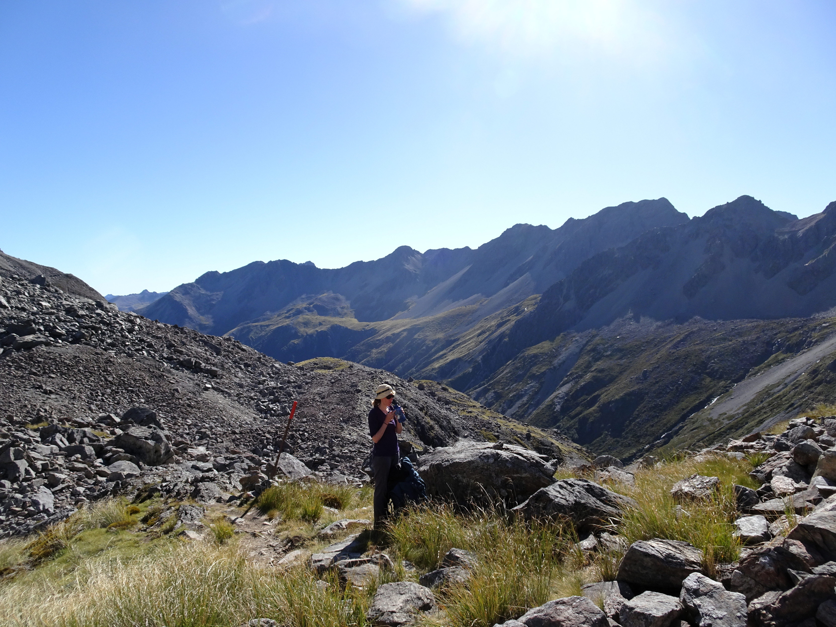 A person taking a water break in the backcountry. There is a marker pole nearby, some scree just behind, and a mountainous ridge in the background, underneath a high sun.