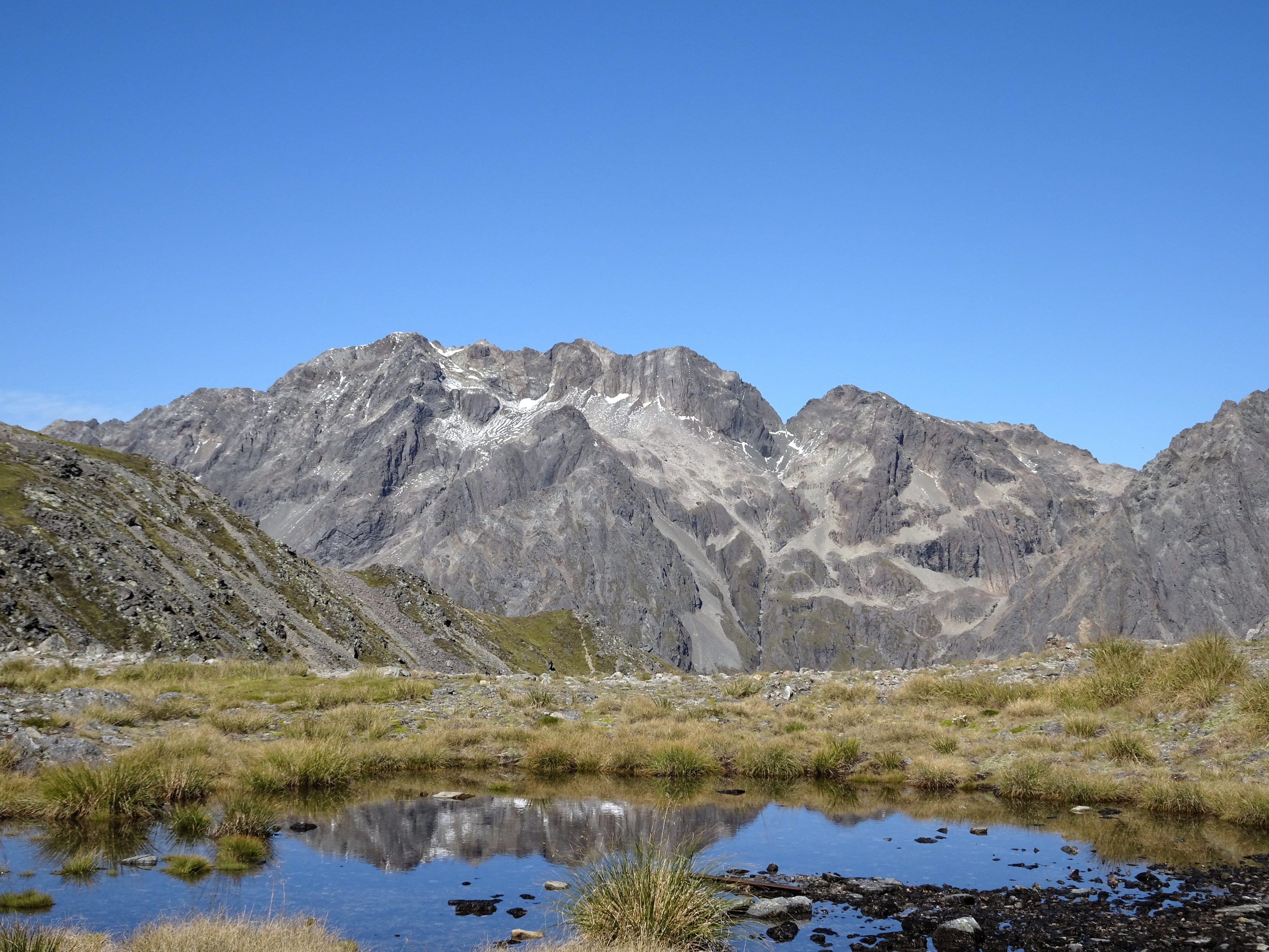 A tarn underneath another mountain ridge. The tarn is reflecting the ridge. There are some rocks and some plants in the tarn.