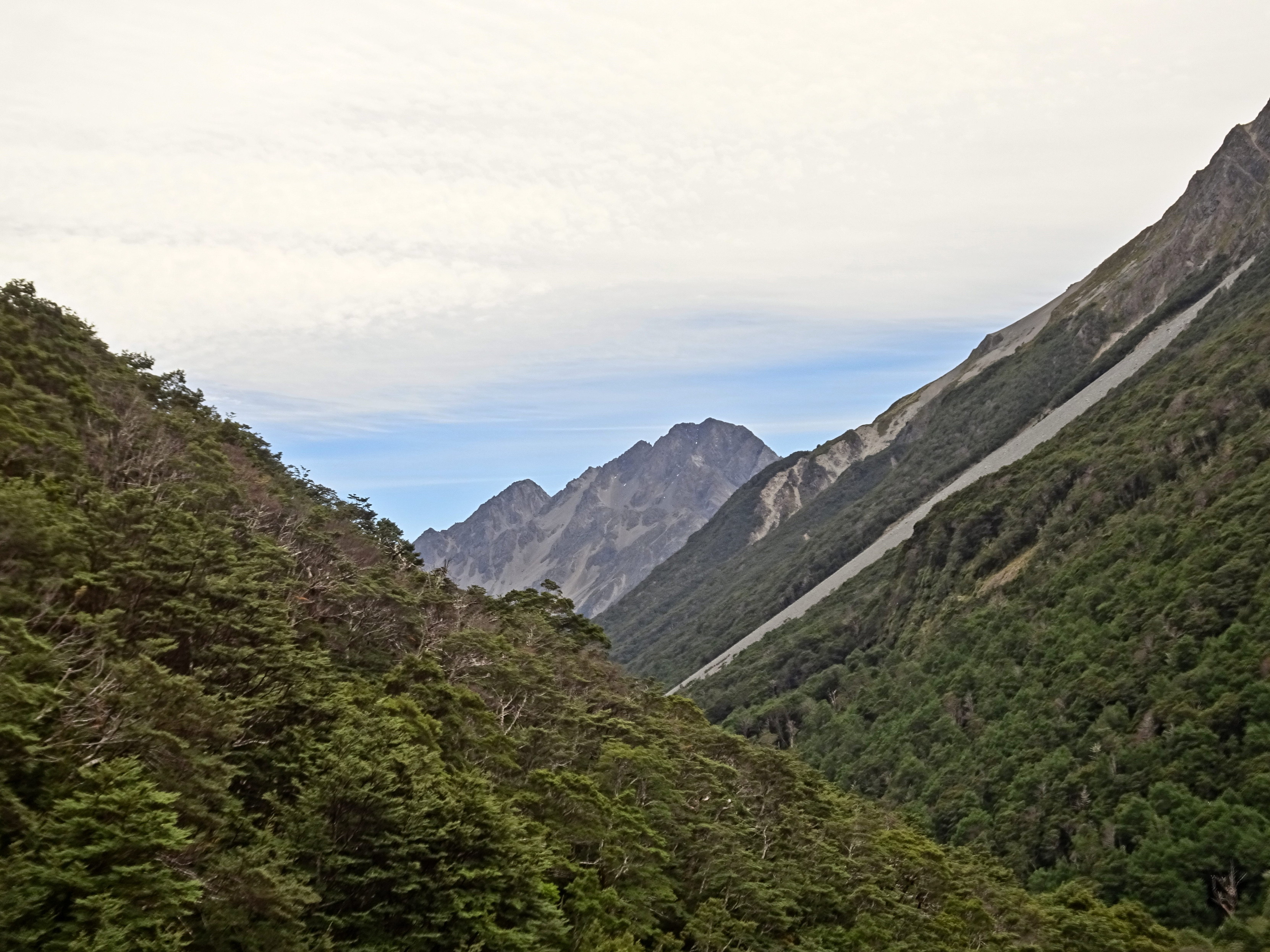Yet another valley picture, with a slope going down and to the right in front, a slope going down and to the left behind it, and two peaks behind. There is high cloud and a bit of blue sky underneath.