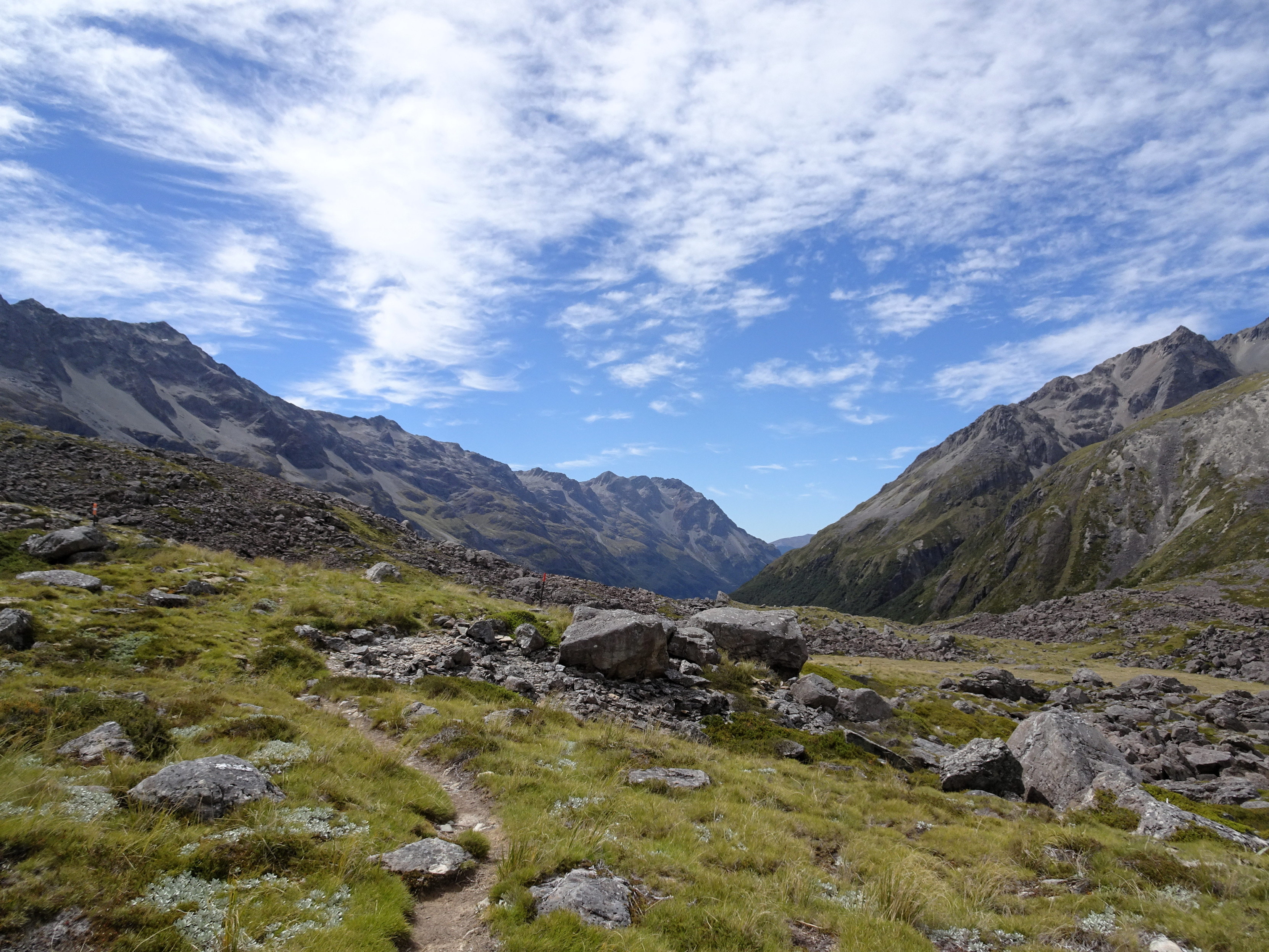 The ground is gently sloping down, with grass and some rocks, and a path through it. More mountains in the background.