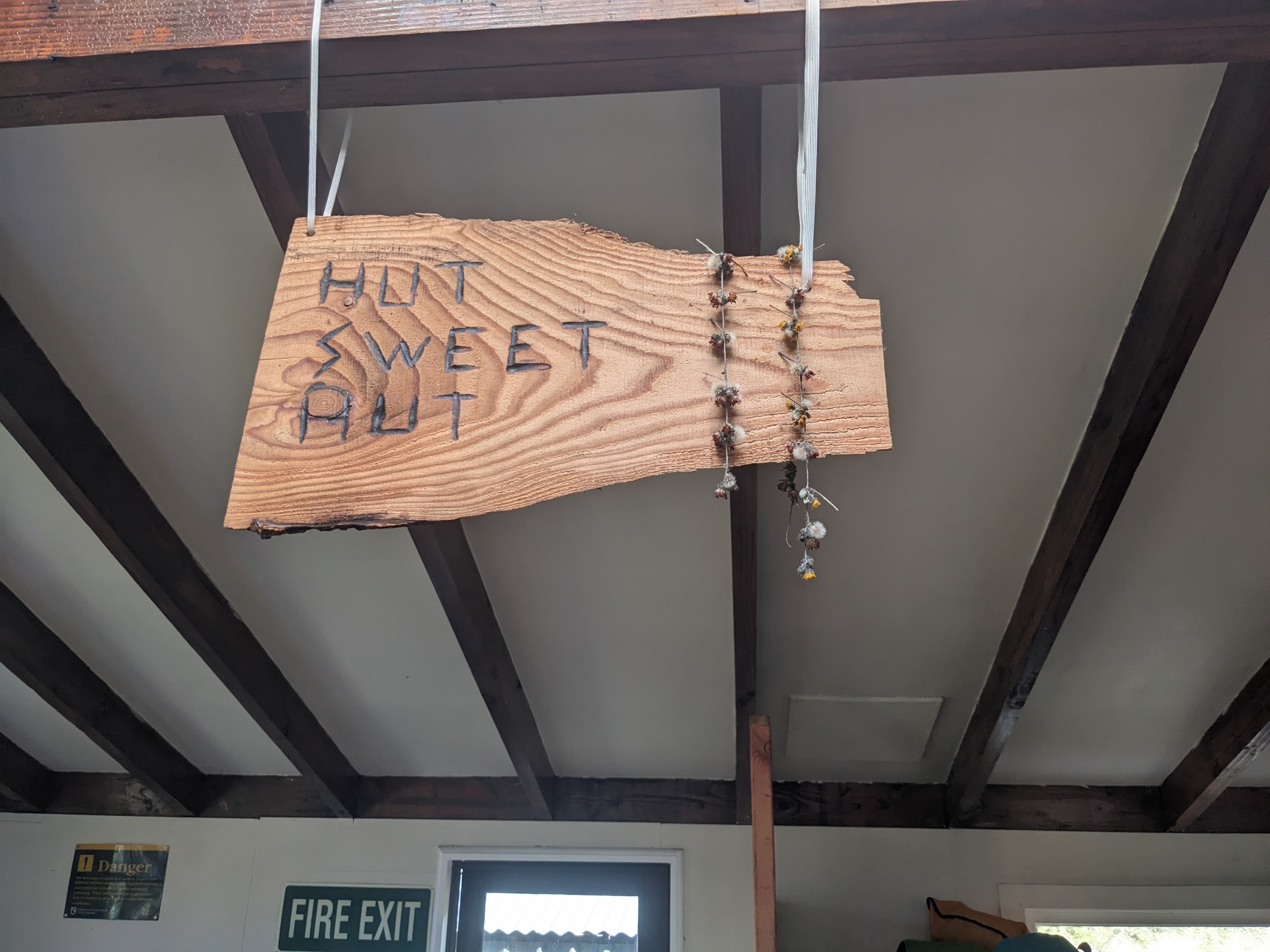 A hanging wooden plank with "Hut Sweet Hut" burned into it, hanging from a rafter. Some flowers tied in a line are hanging off the right-hand side of the plank.