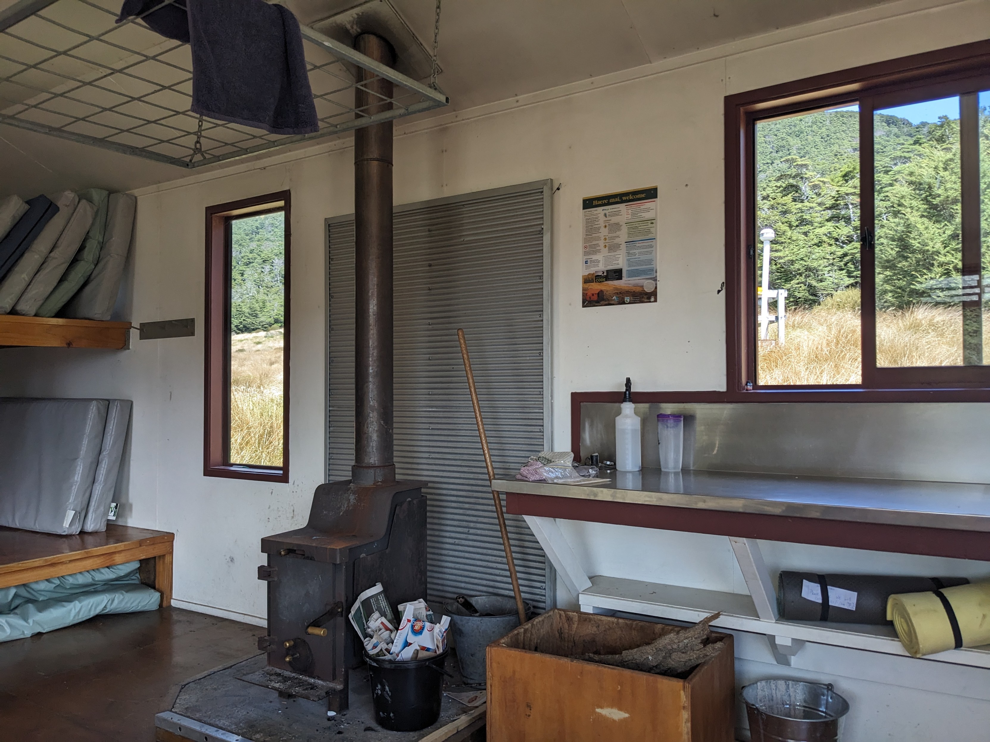 Inside Speargrass Hut. To the right there is a bench for food preparation, with a box for wood underneath it, with a bucket for ash. In the centre there is a wood stove, with metal behind it to prevent flames, and another bucket with paper. There are two reasonable windows. And to the left there are 7 mattresses on two levels, standing on their sides. There is also a mattress underneath the bottom level.