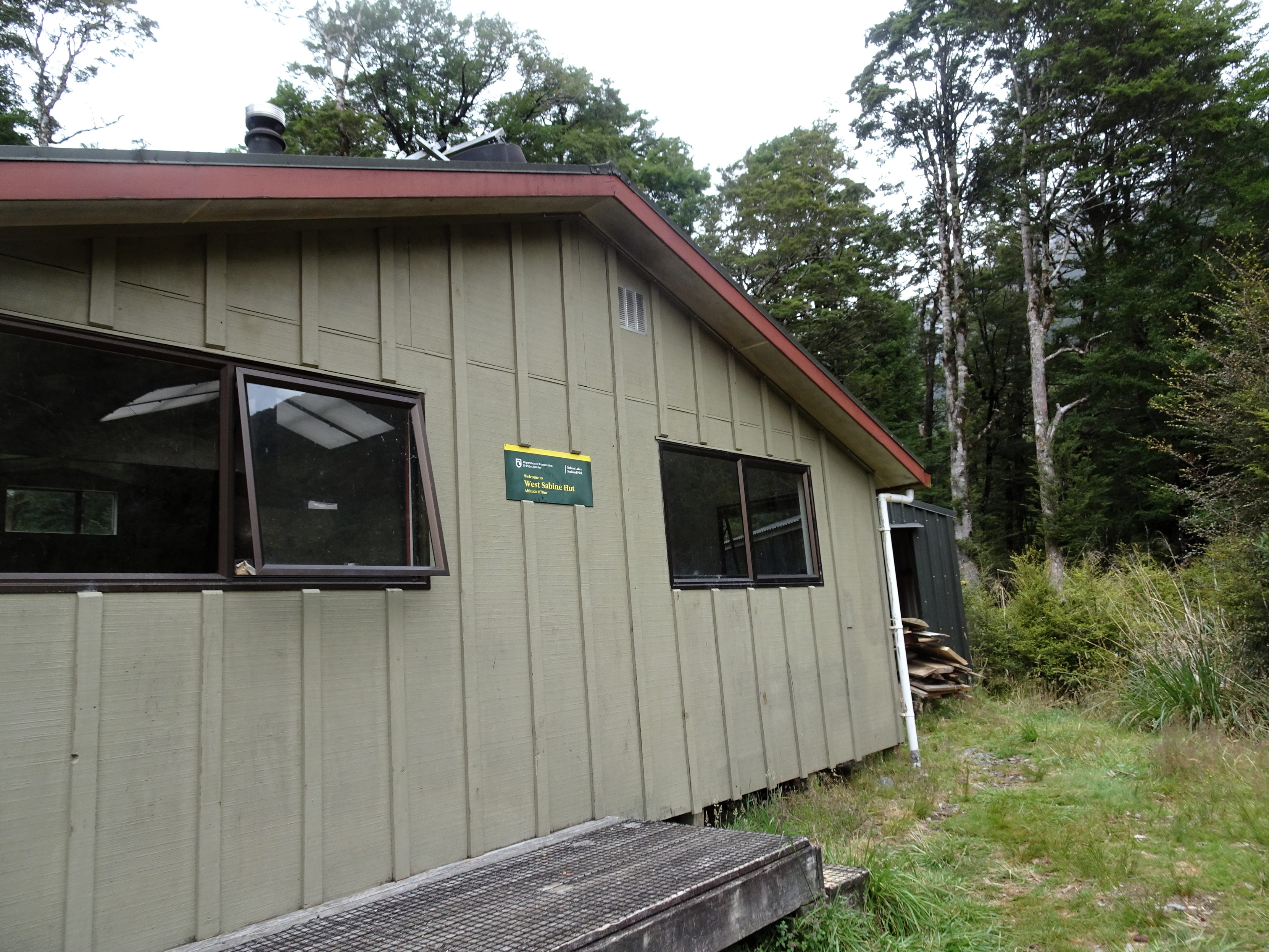 The outside of the West Sabine Hut, with olive cladding and a dark green DOC sign.