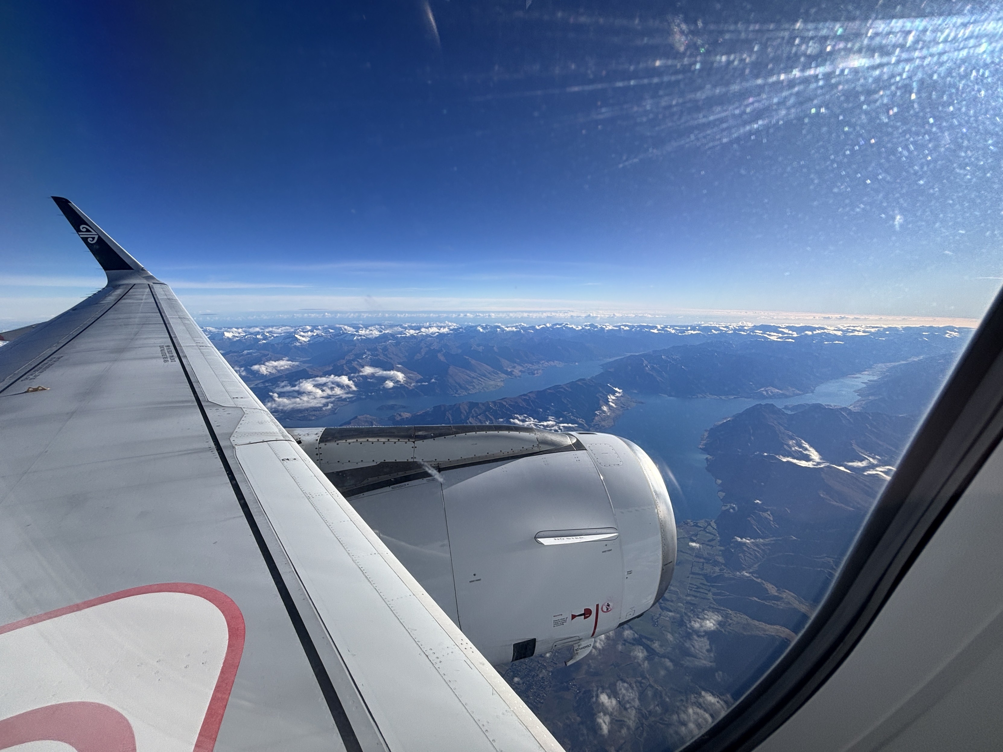 The view out the window from seat 10A in an Air New Zealand Airbus A320 airliner - blue skies, a deep blue lake in a mountainous valley.