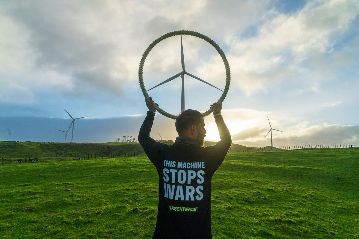 Someone holds a circle up to frame a wind turbine in the distance, making it look like a giant peace sign. They're wearing a t-shirt with the message: "This machine stops wars"