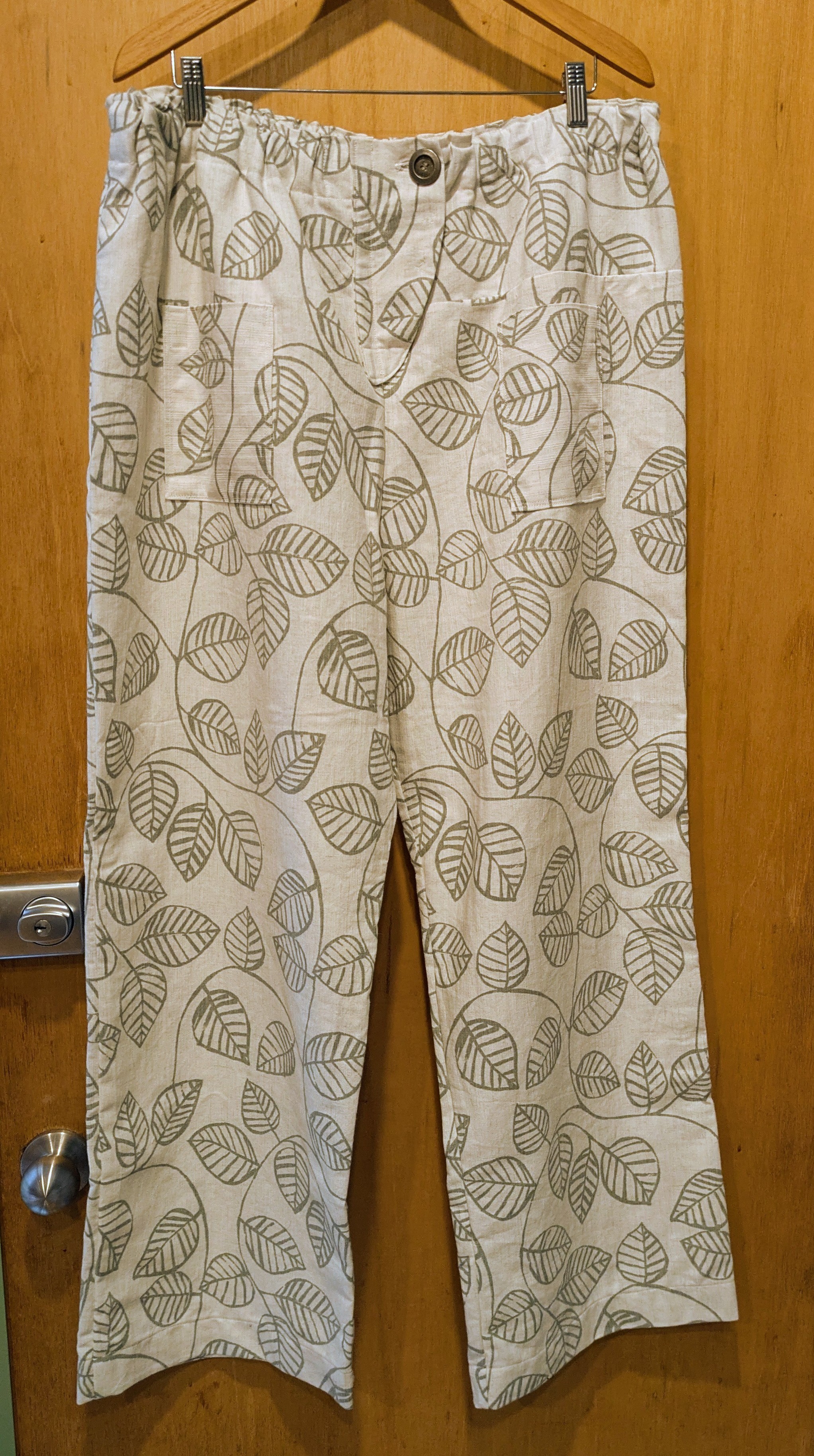 A pair of wide-legged trousers on a hanger. The trousers are off-white with a leaf pattern in grey. The background is a wooden door.