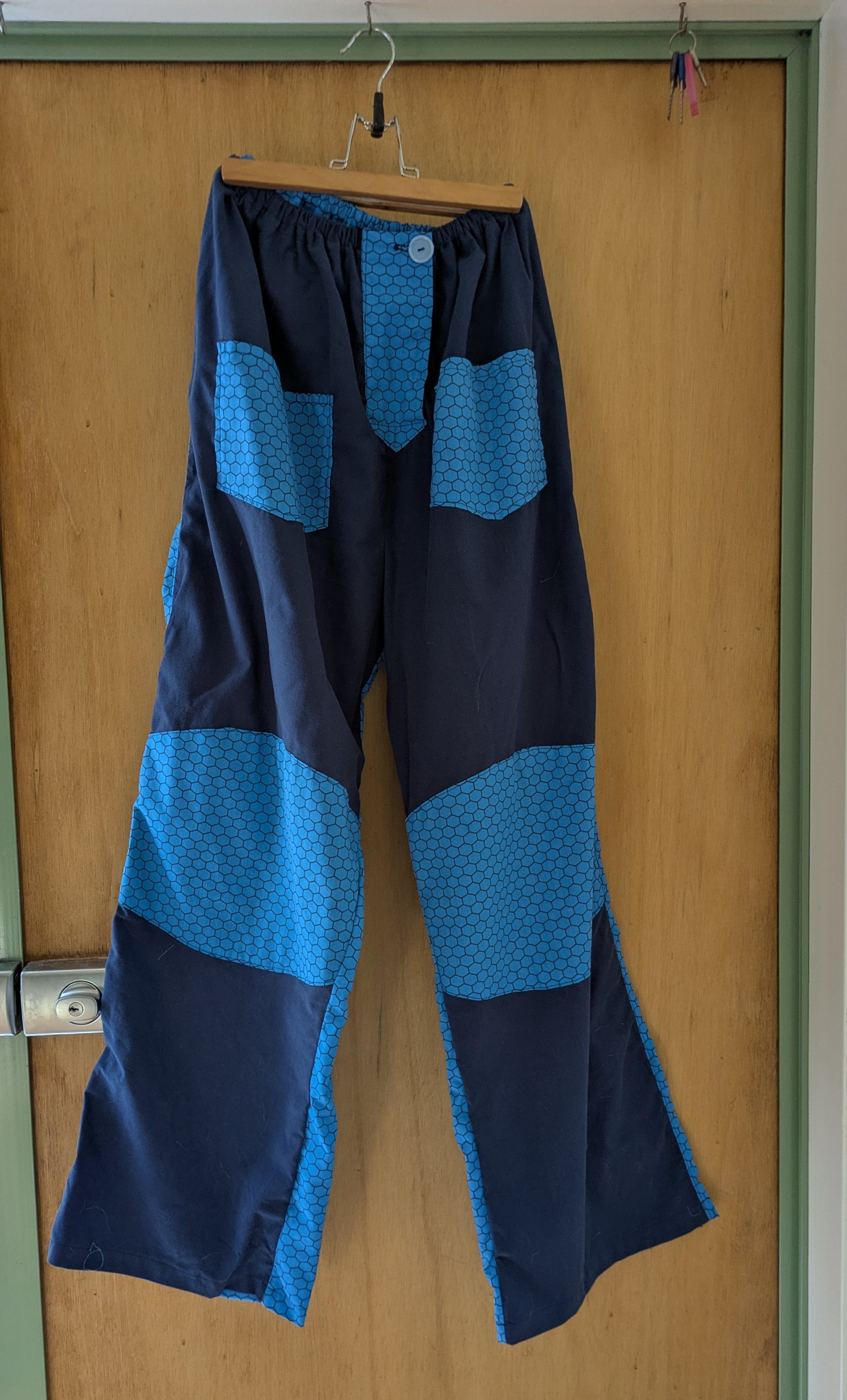 A pair of wide-legged trousers on a hanger. The front of the trousers is dark blue. It has pockets and knee patches in a lighter blue colour with a black honeycomb pattern. The back of the trousers is entirely made of the honeycomb fabric. The background is a wooden door with a pale green frame.