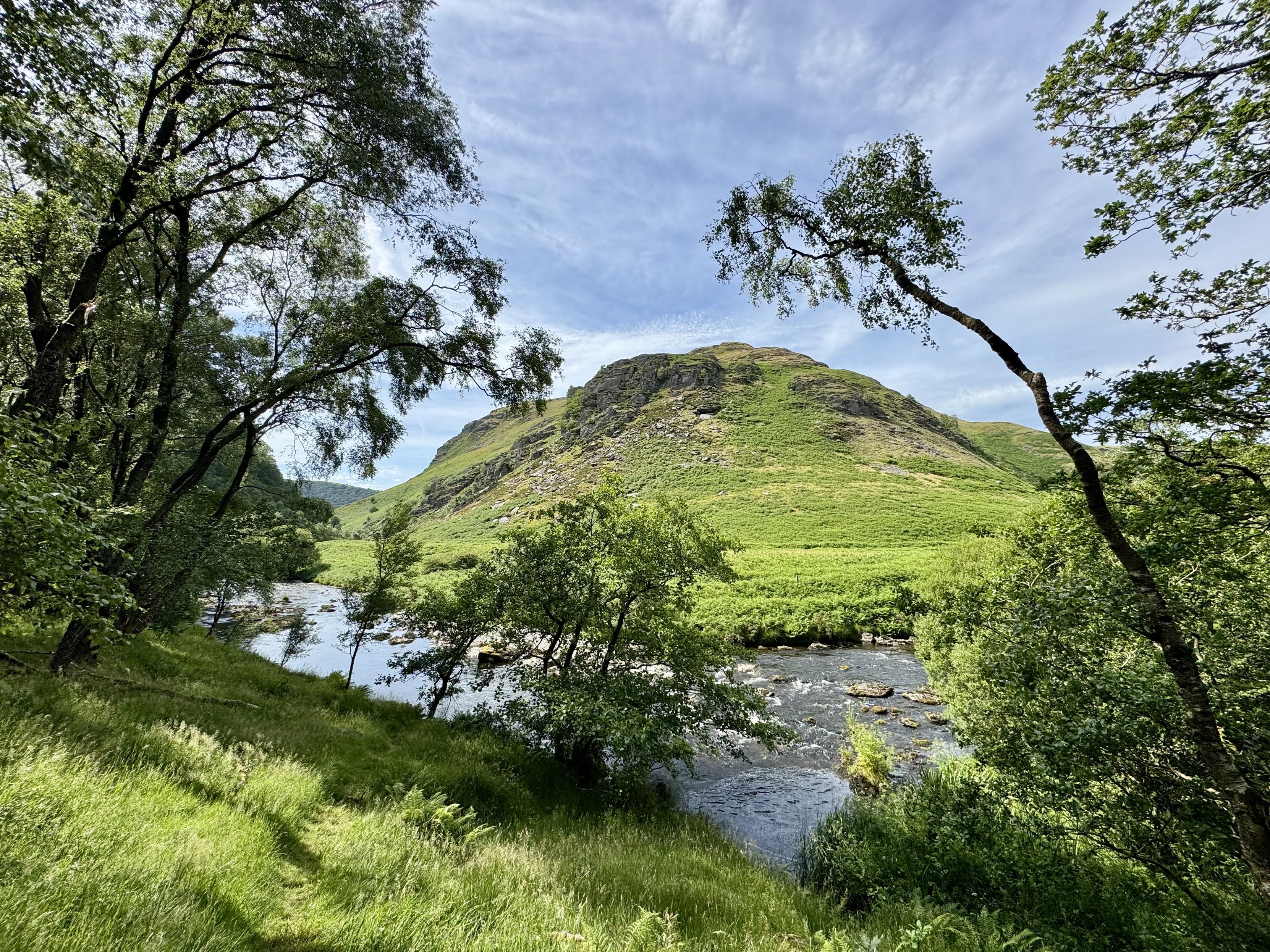 View of clear river from path at the edge of a woodland; bracken-covered hill on opposite bank