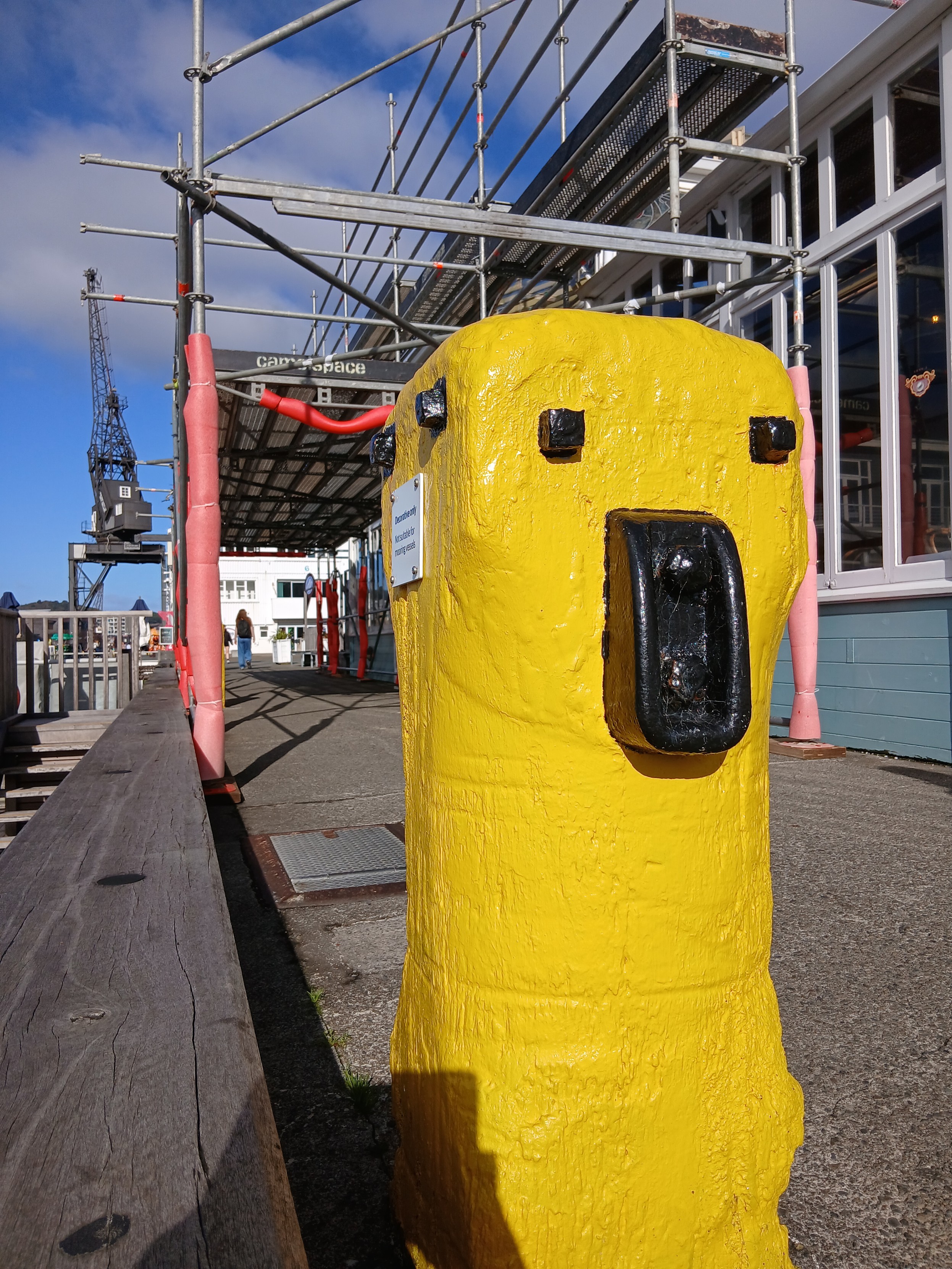 A wooden bollard painted brught yellow sits between a low wooden rail and a building covered in scaffold. Its black bolts and other bits give it a face whuch looks concerned.