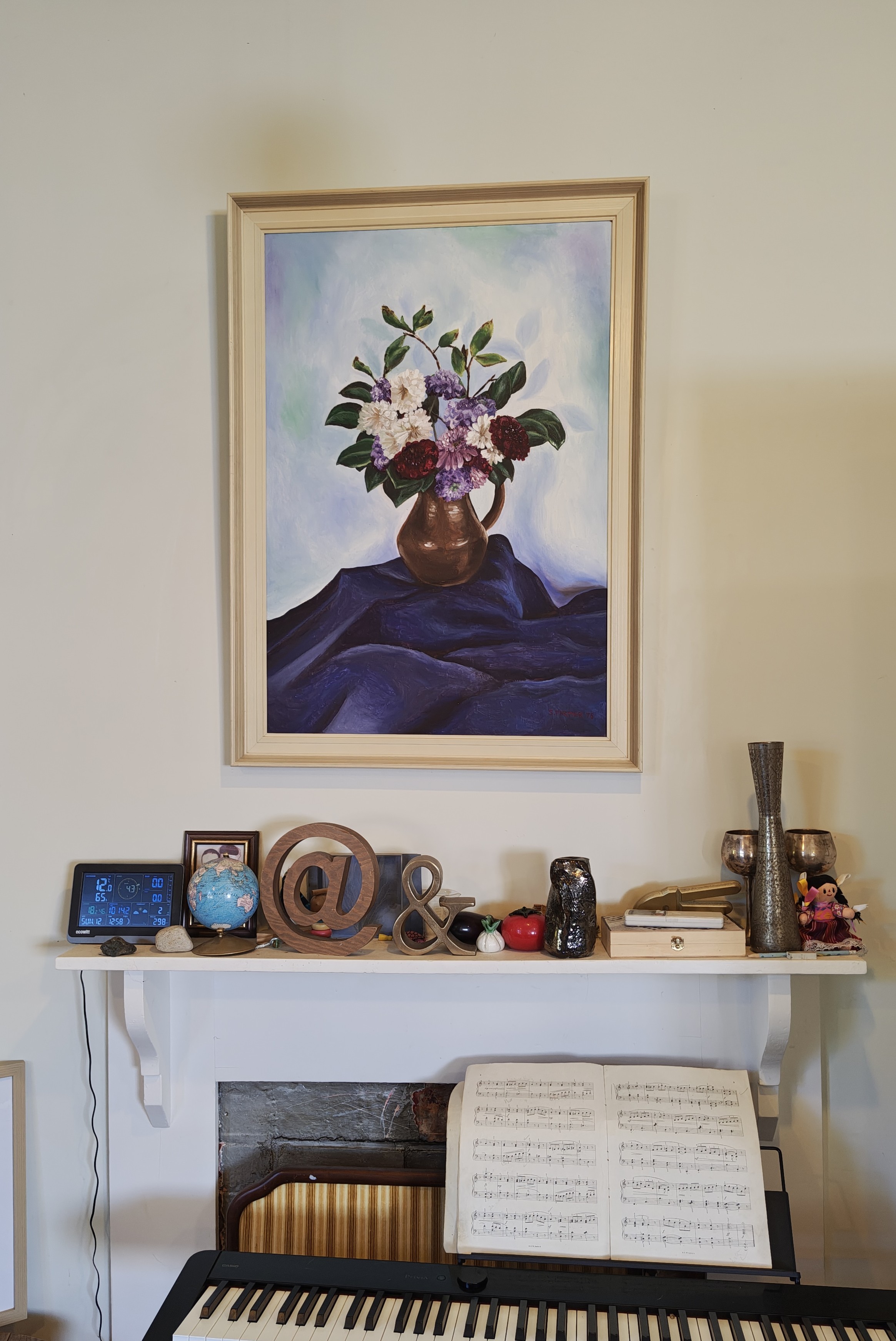 Still life painting of hydrangeas in a vase on a purple fabric covered base. Below the painting is a mantelpiece covered with bric a brac, and below that a piano keyboard.
