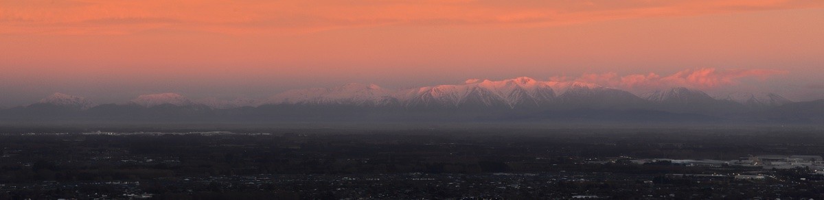 My horizontal panorama photo of snow capped mountains lit up pink by the sunrise, with the farmland and towns of the Canterbury Plains in the foreground.