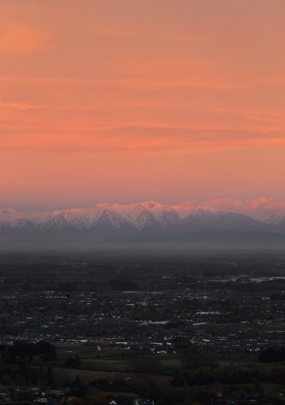 My vertical panorama photo of snow capped mountains lit up pink by the sunrise, with the farmland and towns of the Canterbury Plains, and Christchurch surburbs, in the foreground.