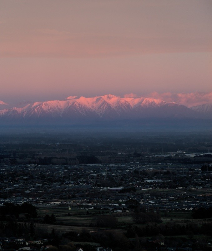 My vertical panorama photo of snow capped mountains lit up pink by the sunrise, with the farmland and towns of the Canterbury Plains, and Christchurch surburbs, in the foreground. This was taken a few minutes later. The clouds are less pink and now there's pink sunlight on the snow of the mountains.