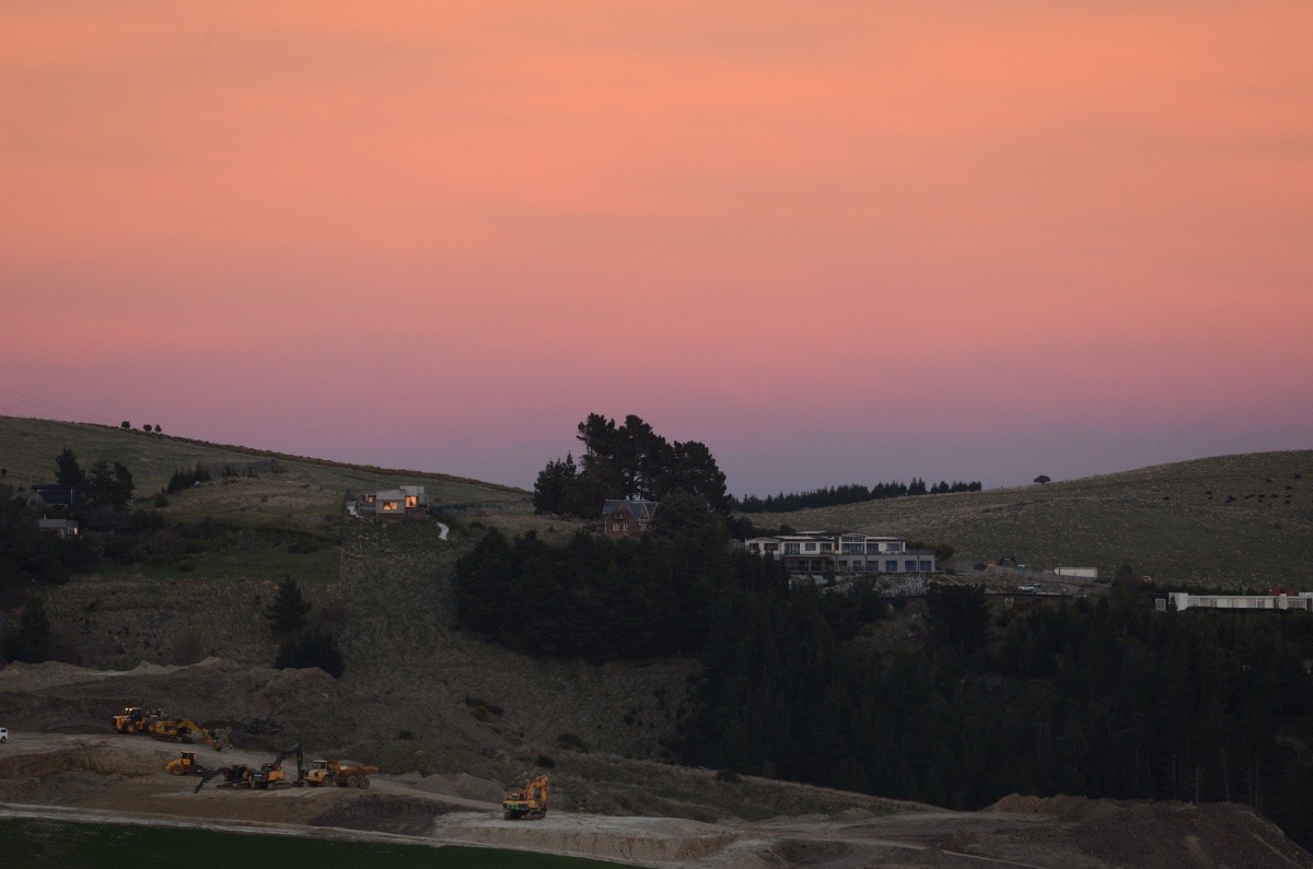 A photo of hills and a few trees and houses with pink clouds behind, at sunrise.
