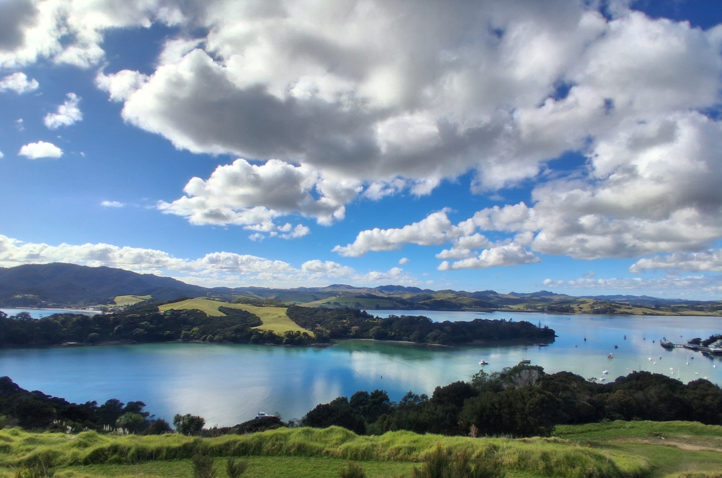 Panorama over a sheltered harbour. Foreground is a grassy slope. Water calm, reflecting sky. Blue hills in the background. Sky blue with puffy white clouds.
