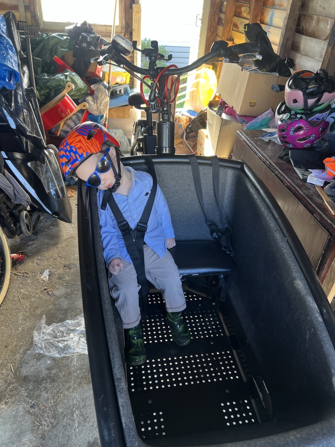 A young child wearing a colorful helmet and sunglasses is sleeping in a bike cargo compartment. The bike is parked in a garage surrounded with various items, including other bikes and helmets. Through the other end of the garage, you can see it is a bright sunny day.