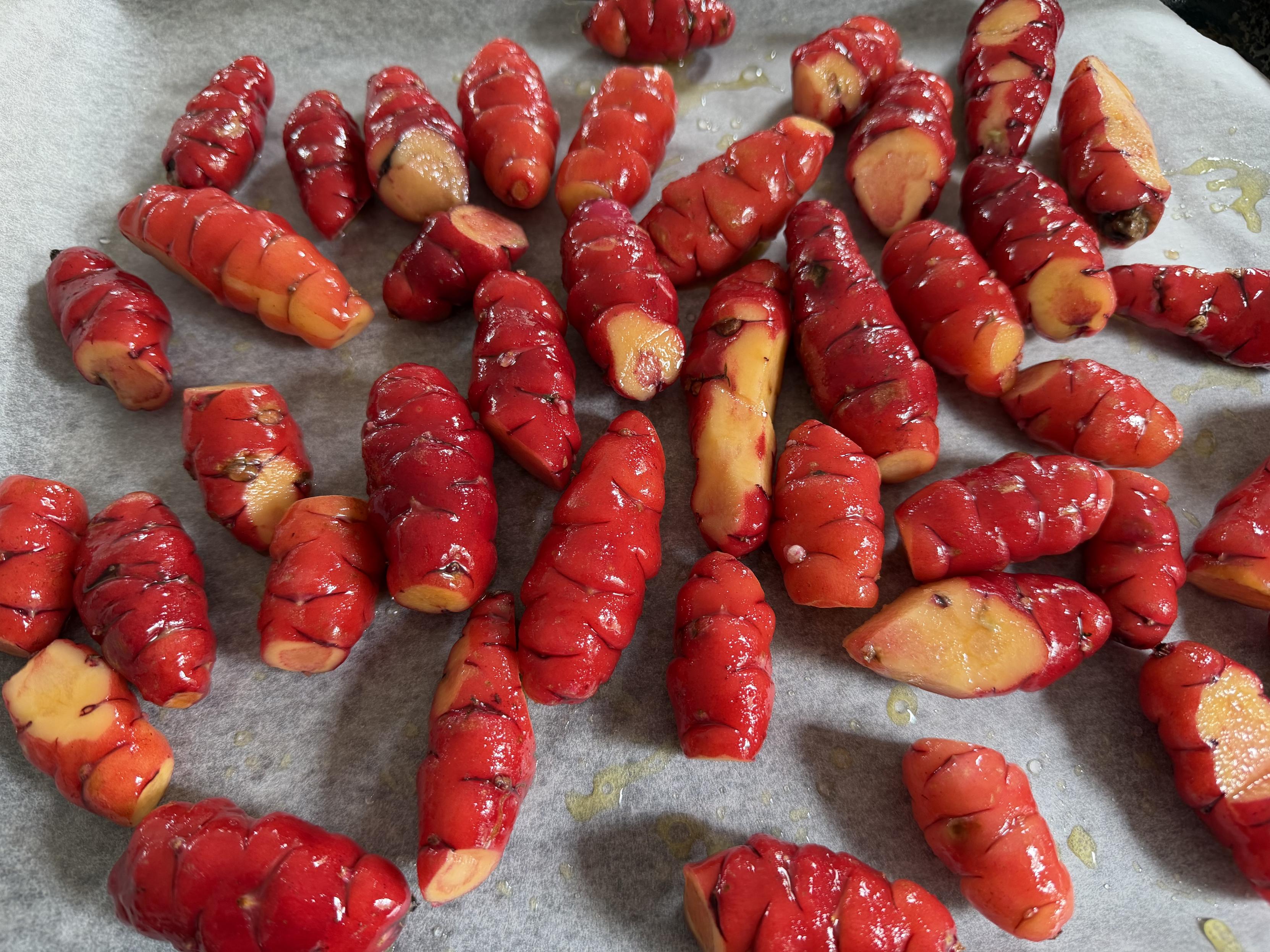 This image features a scattered arrangement of vibrant red, glossy root vegetables, likely New Zealand yams (oca), spread across a piece of parchment paper. The tubers have a distinct, segmented, and slightly wrinkled texture, with some cut open to reveal a bright yellow-orange interior. A light sheen of oil is visible on the vegetables and the paper, suggesting they are prepared for roasting.